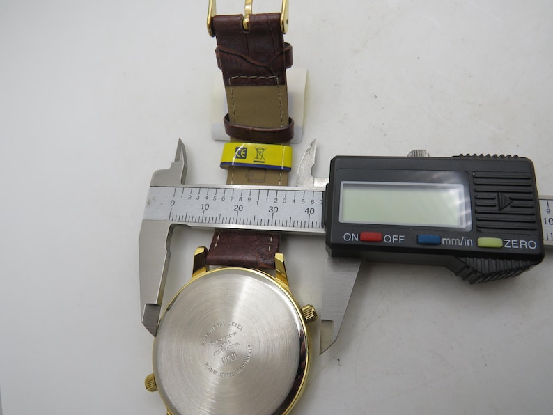 May include: A gold-toned wristwatch with a brown leather band is measured with a digital caliper. The watch face is visible, and the band is partially unclasped. The caliper displays a measurement in millimeters and inches.