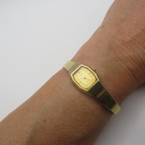 May include: A gold wristwatch with an octagonal face and a matching band. The watch features a gold dial with black hands and markers. The band combines gold metal and a cream-coloured material.