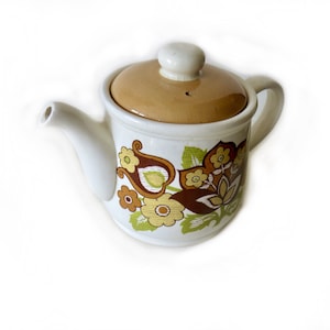 May include: A white ceramic teapot with a brown lid and a floral pattern in shades of brown, yellow, and green.