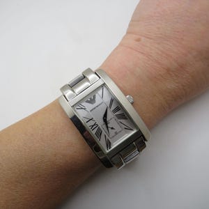 Puede incluir: Un reloj plateado con una esfera rectangular y una correa plateada. La esfera del reloj tiene números romanos y el nombre de la marca "Emporio Armani" impreso en ella.