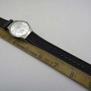 May include: A silver wristwatch with a white face and black rubber band. The watch is laid on a wooden ruler with markings in inches.