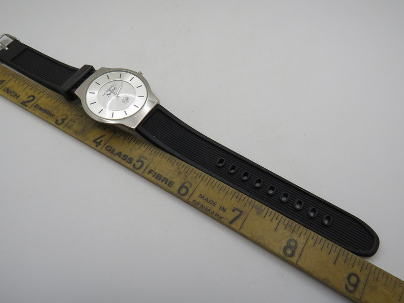 May include: A silver wristwatch with a white face and black rubber band. The watch is laid on a wooden ruler with markings in inches.