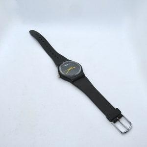 K&ouml;nnte beinhalten: Schwarze Armbanduhr mit rundem Zifferblatt und gelbem Zeiger. Die Uhr hat ein schwarzes Armband mit silberner Schnalle. Das Zifferblatt hat einen gelben Zeiger und einen schwarzen Hintergrund.