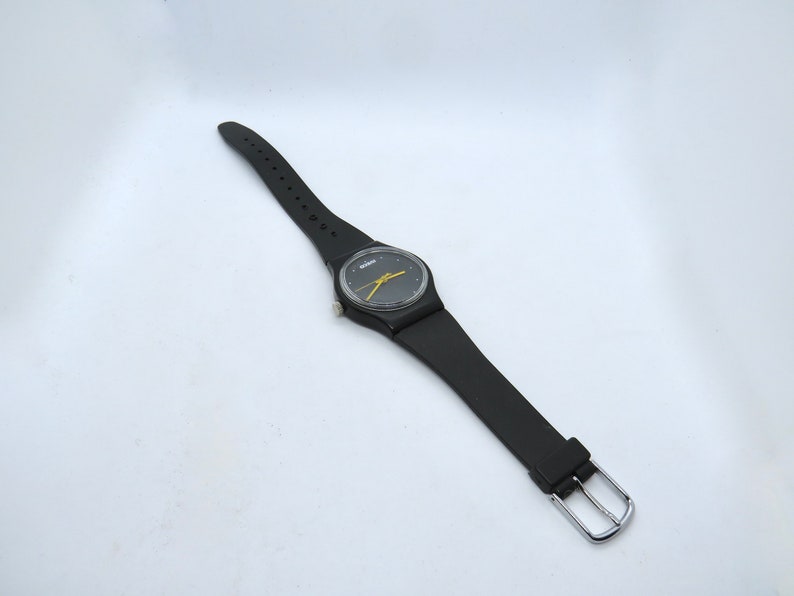 K&ouml;nnte beinhalten: Schwarze Armbanduhr mit rundem Zifferblatt und gelbem Zeiger. Die Uhr hat ein schwarzes Armband mit silberner Schnalle. Das Zifferblatt hat einen gelben Zeiger und einen schwarzen Hintergrund.