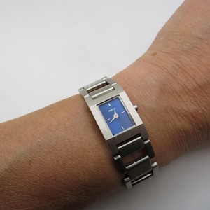 Puede incluir: Un reloj plateado con una esfera azul rectangular. El reloj tiene una correa de acero inoxidable y la palabra "MOXX" está impresa en la esfera. El reloj está en una muñeca.