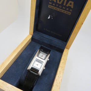 Puede incluir: Un reloj Avia plateado con dos esferas cuadradas y una correa de cuero negro, presentado en una caja de madera con interior de terciopelo azul. La tapa de la caja muestra el logotipo de Avia Watches y el texto "Stand the test of time."