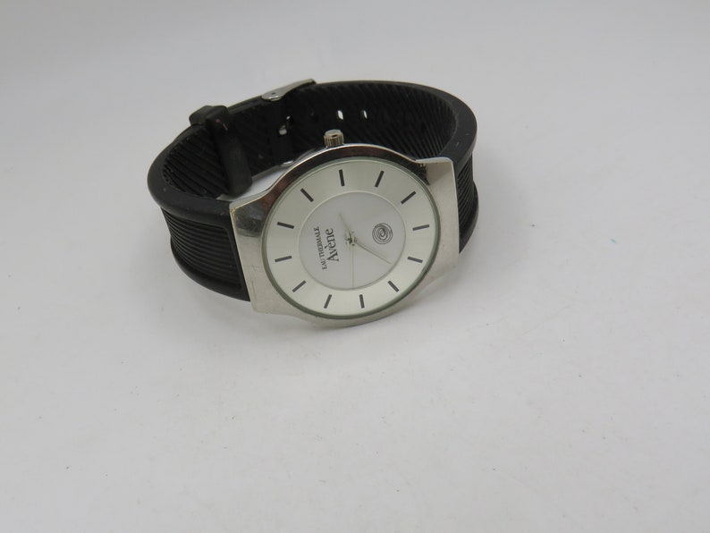 May include: A silver wristwatch with a white face and black rubber band. The watch face has black markings and the word "Avene" printed on it.