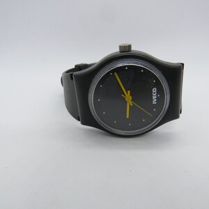 K&ouml;nnte beinhalten: Schwarze Armbanduhr mit rundem schwarzem Zifferblatt und gelben Zeigern. Die Uhr hat ein schwarzes Armband und eine silberne Krone. Auf dem Zifferblatt steht das Wort "IVECO" in Wei&szlig;. Auf wei&szlig;em Hintergrund.