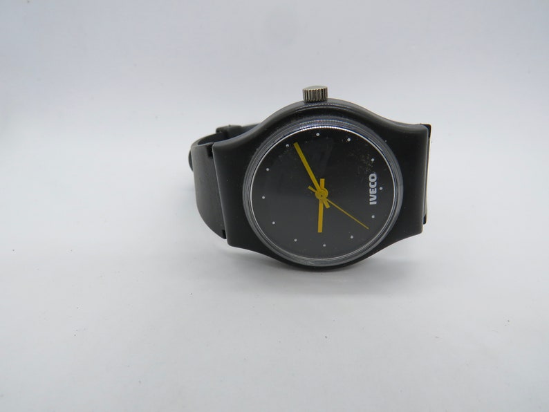 K&ouml;nnte beinhalten: Schwarze Armbanduhr mit rundem schwarzem Zifferblatt und gelben Zeigern. Die Uhr hat ein schwarzes Armband und eine silberne Krone. Auf dem Zifferblatt steht das Wort "IVECO" in Wei&szlig;. Auf wei&szlig;em Hintergrund.