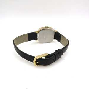 K&ouml;nnte beinhalten: Eine goldfarbene Uhr mit quadratischem Zifferblatt und schwarzem Lederarmband. Die Uhr hat ein silbernes Zifferblatt mit schwarzen Markierungen.