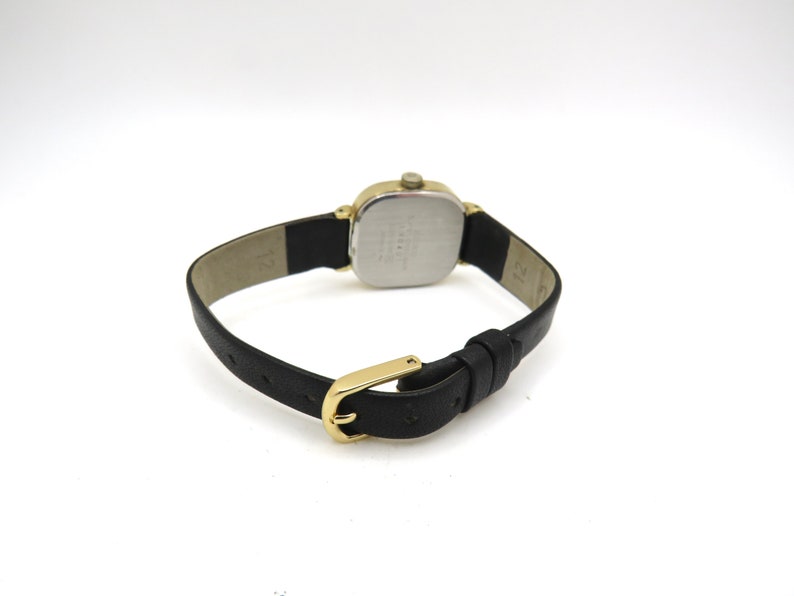 K&ouml;nnte beinhalten: Eine goldfarbene Uhr mit quadratischem Zifferblatt und schwarzem Lederarmband. Die Uhr hat ein silbernes Zifferblatt mit schwarzen Markierungen.