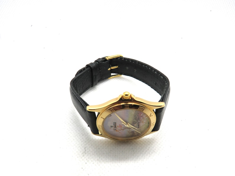 May include: A gold-tone wristwatch with a black leather band. The watch face has a white background with a floral design and Roman numeral hour markers. The watch face also has the word "Zodiac" printed on it.
