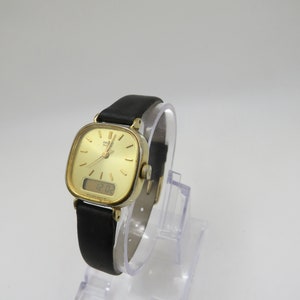 K&ouml;nnte beinhalten: Eine goldfarbene analog-digitale Armbanduhr mit einem schwarzen Lederarmband. Das Zifferblatt der Uhr ist quadratisch und hat einen wei&szlig;en Hintergrund mit schwarzen Markierungen. Die digitale Anzeige zeigt die Zeit 12:03 an.