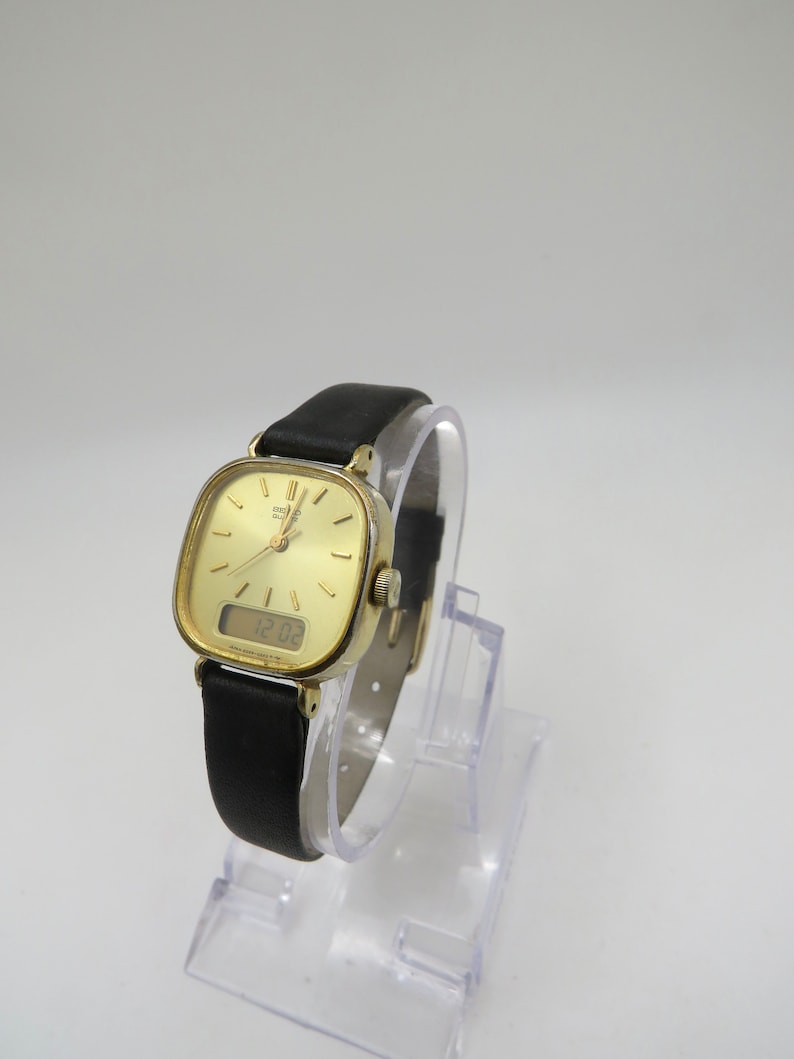 K&ouml;nnte beinhalten: Eine goldfarbene analog-digitale Armbanduhr mit einem schwarzen Lederarmband. Das Zifferblatt der Uhr ist quadratisch und hat einen wei&szlig;en Hintergrund mit schwarzen Markierungen. Die digitale Anzeige zeigt die Zeit 12:03 an.