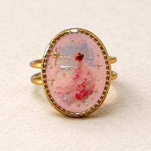 Può includere: Un anello ovale dorato con una scena dipinta in rosa raffigurante una figura con un abito fluido e accenti floreali rossi. Il design include piccoli dettagli a forma di stella e un bordo dorato testurizzato. Il testo @SantomaroDesign è sopra.