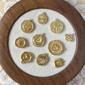 Könnte beinhalten: Eine Sammlung goldfarbener Himmelscharms, darunter Sonnen, Monde und Sterne, angeordnet in einem Holzrahmen. Die Charms weisen aufwendige Details auf und werden auf weißem Hintergrund präsentiert. Das Bild enthält den Text "@SantamaroDesign".