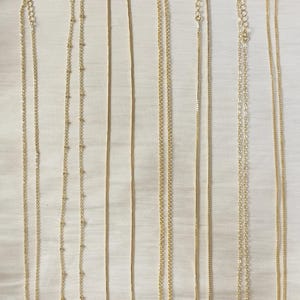 Op de afbeelding: Een verzameling delicate gouden kettingen, gepresenteerd op een witte achtergrond. De kettingen vari&euml;ren in stijl, waaronder ketting-, kralen- en slangenkettingontwerpen. Elke ketting heeft een sluiting en een verlengketting. De tekst "@SantomaroDesign" is inbegrepen.
