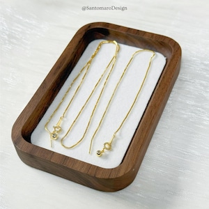 May include: A gold-colored necklace and earrings set displayed in a wooden tray with a white interior. The necklace has a small pendant, and the earrings are threader style. The text "@SantamaroDesign" is in the top left corner.