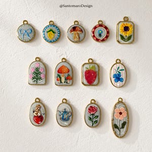 Pode incluir: Uma coleção de doze pingentes dourados com vários designs coloridos. Os pingentes apresentam imagens de um laço, limão, cogumelo, flor, girassol, morango, anjo, bule, rosa e outros motivos florais e frutados.