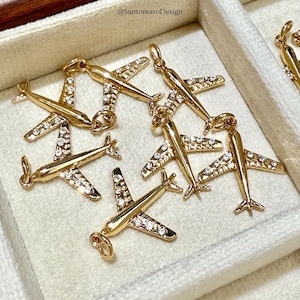 Peut inclure: Charm en forme d'avion de couleur or, orné de cristaux transparents. Les breloques sont disposées dans un écrin à bijoux blanc. Chaque avion est doté d'une boucle pour l'attacher à un collier ou un bracelet. L'image est bien éclairée.