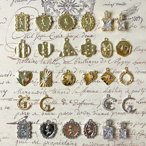Puede incluir: Una colección de colgantes y abalorios dorados en varias formas, incluyendo corazones, círculos y rectángulos. Algunos presentan diseños florales, mientras que otros tienen patrones geométricos o piedras rectangulares transparentes. El fondo es papel de estilo vintage.
