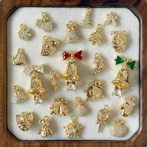 Pode incluir: Uma coleção de pingentes dourados com tema natalino, incluindo sinos, flocos de neve, árvores de Natal, laços e um boneco de gengibre. Alguns pingentes são adornados com cristais transparentes, em uma bandeja de madeira.