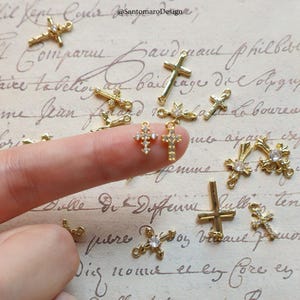 May include: A collection of gold-colored cross charms, some adorned with clear gemstones. The charms vary in design, with some featuring a simple cross shape and others with intricate detailing. The background is a vintage-style paper with handwritten script.