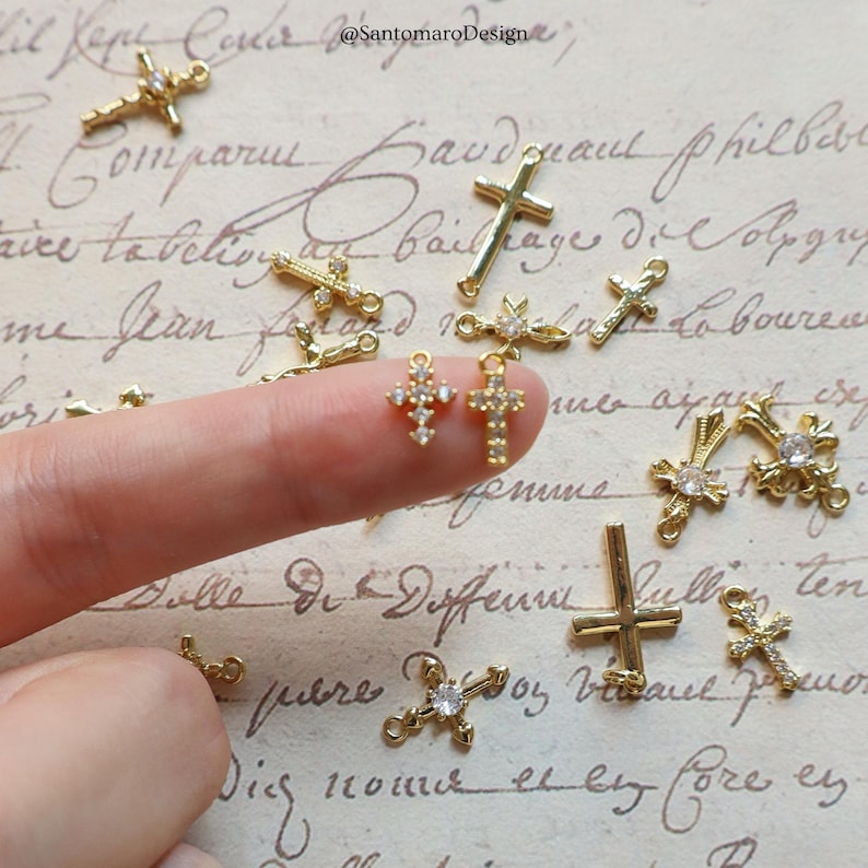 May include: A collection of gold-colored cross charms, some adorned with clear gemstones. The charms vary in design, with some featuring a simple cross shape and others with intricate detailing. The background is a vintage-style paper with handwritten script.