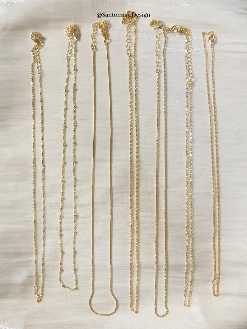 Op de afbeelding: Een verzameling delicate gouden kettingen, gepresenteerd op een witte achtergrond. De kettingen vari&euml;ren in stijl, waaronder ketting-, kralen- en slangenkettingontwerpen. Elke ketting heeft een sluiting en een verlengketting. De tekst "@SantomaroDesign" is inbegrepen.