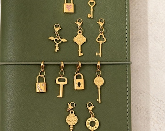 Könnte beinhalten: Goldfarbene Anhänger in verschiedenen Formen, darunter Schlüssel und Vorhängeschlösser, werden vor einem grünen Hintergrund präsentiert. Die Anhänger weisen aufwendige Designs wie Herzen, Blumen und geometrische Muster auf. Die Anhänger sind an einer Schnur befestigt.