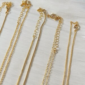 Op de afbeelding: Close-up van verschillende goudkleurige kettingen met verschillende kettingstijlen. Elke ketting heeft een sluiting en een verstelbare ketting. De achtergrond is een witte, getextureerde stof. De tekst "@SantomaroDesign" is zichtbaar.