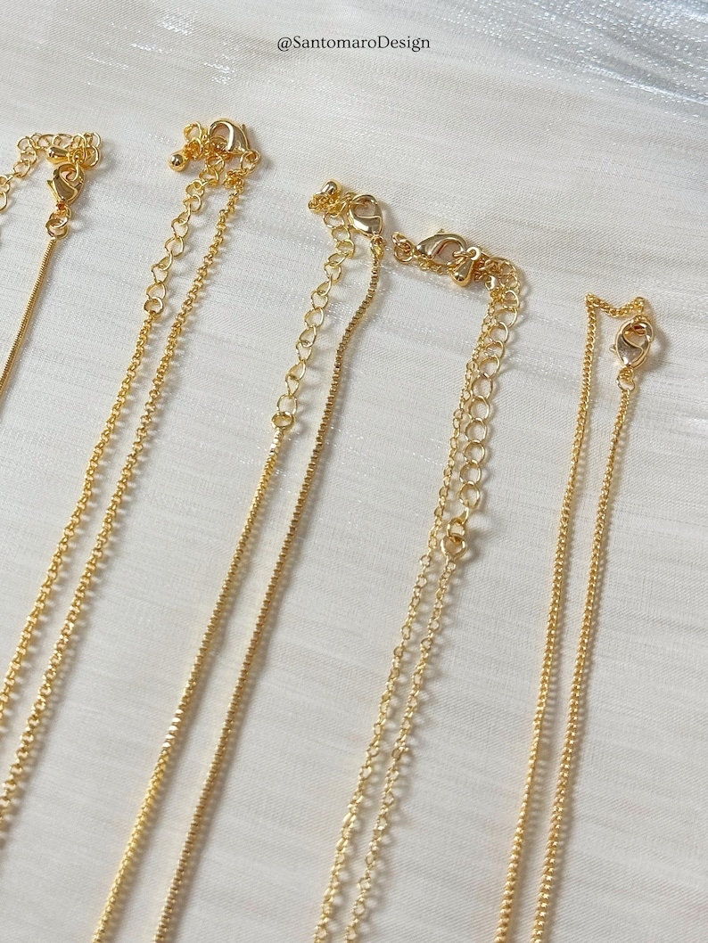 Op de afbeelding: Close-up van verschillende goudkleurige kettingen met verschillende kettingstijlen. Elke ketting heeft een sluiting en een verstelbare ketting. De achtergrond is een witte, getextureerde stof. De tekst "@SantomaroDesign" is zichtbaar.