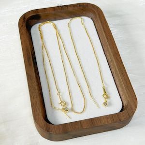 May include: Gold-colored threader earrings displayed in a wooden tray with a white interior. The earrings have a delicate chain design with a small, round charm at the end of each threader. The earrings are presented on a white surface.