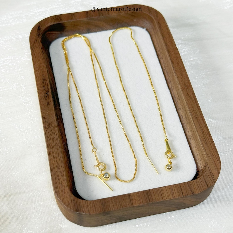 May include: Gold-colored threader earrings displayed in a wooden tray with a white interior. The earrings have a delicate chain design with a small, round charm at the end of each threader. The earrings are presented on a white surface.