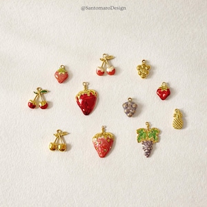 May include: A collection of gold-toned fruit charms, including cherries, strawberries, grapes, and a pineapple. Each charm is detailed with red, purple, and green enamel, and has a small loop for attaching to jewelry or other crafts. The text "@SantomaroDesign" is in the top left.