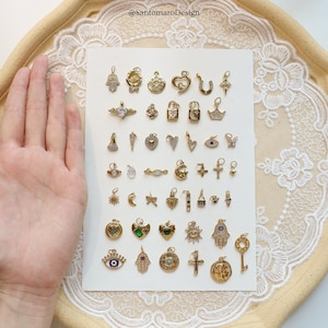May include: A collection of gold-colored charms in various shapes, including hearts, crosses, and keys. Some charms feature gemstones and intricate designs. The charms are displayed on a white surface with a lace trim.