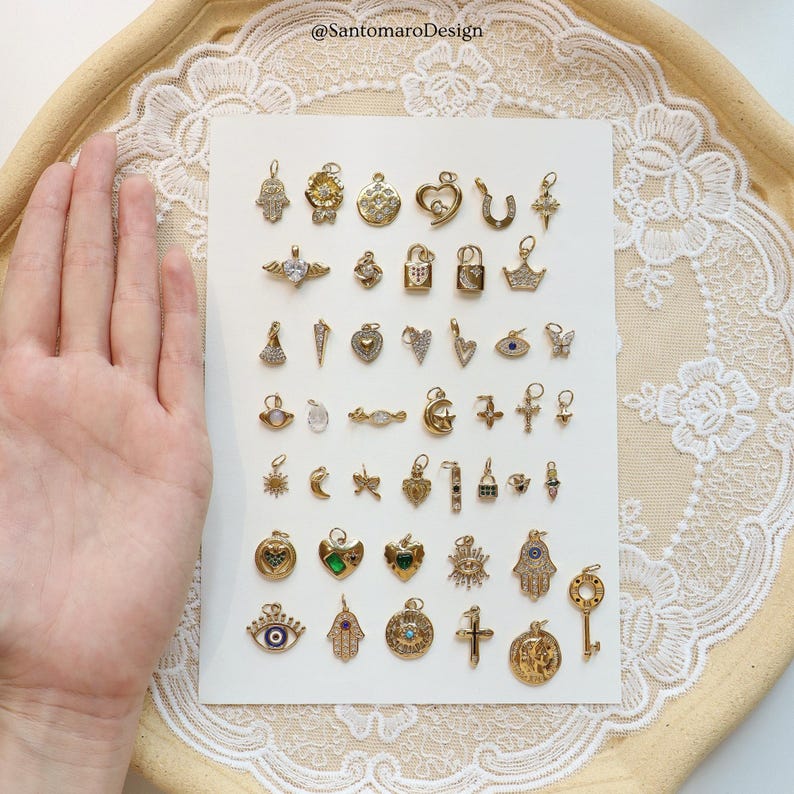 May include: A collection of gold-colored charms in various shapes, including hearts, crosses, and keys. Some charms feature gemstones and intricate designs. The charms are displayed on a white surface with a lace trim.