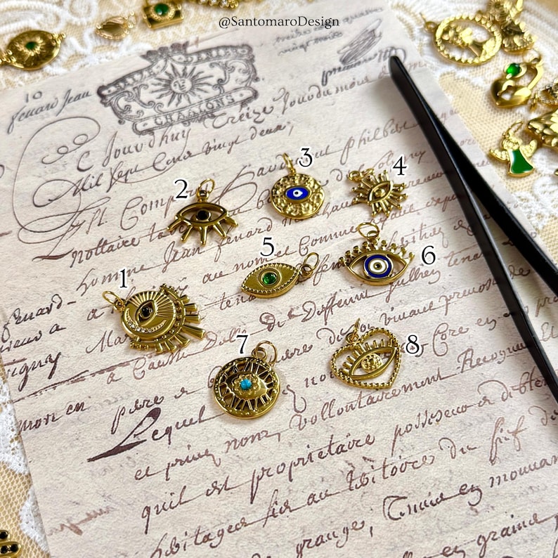 May include: Gold-coloured charm pendants with eye and sun designs. The charms feature intricate details, including blue and green accents. The pendants are displayed on a vintage paper background, showcasing a variety of styles.