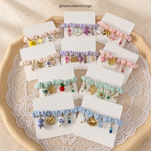 Könnte beinhalten: Eine Sammlung von Scrunchie-Armbändern in verschiedenen Farben, die jeweils mit einzigartigen Charms verziert sind. Die Armbänder verfügen über Charms wie Smiley-Gesichter, Herzen, Sterne und andere dekorative Elemente. Die Scrunchies werden auf weißen Karten präsentiert.