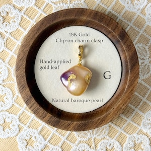May include: A natural baroque pearl pendant with hand-applied gold leaf, attached to an 18K gold clip-on charm clasp. The pearl has a distinctive shape and a mix of colours, including purple and gold. The pendant is displayed in a wooden frame.