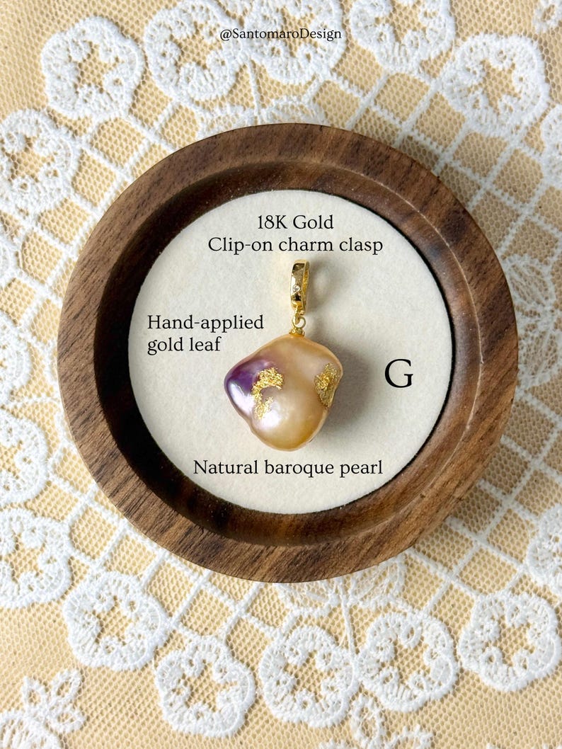 May include: A natural baroque pearl pendant with hand-applied gold leaf, attached to an 18K gold clip-on charm clasp. The pearl has a distinctive shape and a mix of colours, including purple and gold. The pendant is displayed in a wooden frame.