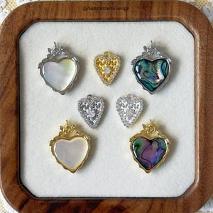 May include: A collection of heart-shaped pendants in various designs. Some feature mother-of-pearl, abalone, or clear stones, set in silver or gold-toned metal frames. The pendants are displayed on a white surface within a wooden tray.