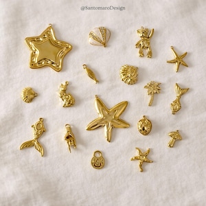 Op de afbeelding: Een verzameling goudkleurige bedels in verschillende vormen, waaronder sterren, schelpen, een teddybeer, een kat, een vis, een hand, een slot, een palmboom, een zeemeerminstaart en een paddenstoel. De bedels zijn op een wit oppervlak geplaatst.
