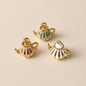 May include: Three miniature teapot charms featuring a gold-tone metal finish with vertical stripes in light blue, soft pink, and white enamel. Each charm includes a small loop at the top for attachment and displays the text @SantomaroDesign above them.