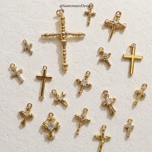 May include: A collection of gold-colored cross charms in various designs. Some crosses are adorned with clear gemstones, while others have a plain, polished finish. The charms are arranged on a textured, off-white surface.