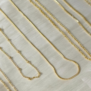 Op de afbeelding: Een verzameling delicate gouden kettingen in verschillende stijlen, waaronder een slangenketting, kralenkettingen en schakelkettingen. De kettingen zijn op een wit oppervlak geplaatst, waarbij hun verschillende ontwerpen en texturen worden getoond. De afbeelding is goed verlicht en benadrukt de glans van het goud.