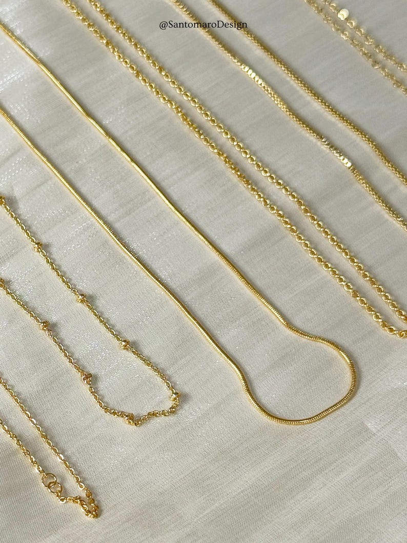 Op de afbeelding: Een verzameling delicate gouden kettingen in verschillende stijlen, waaronder een slangenketting, kralenkettingen en schakelkettingen. De kettingen zijn op een wit oppervlak geplaatst, waarbij hun verschillende ontwerpen en texturen worden getoond. De afbeelding is goed verlicht en benadrukt de glans van het goud.