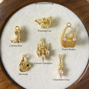 May include: A collection of gold-colored charms, including a cowboy boot, watering can, flower basket, crystal chandelier, mermaid, and chandelier drop. The charms are displayed on a white surface within a wooden frame. Text on the image reads "@SantomaroDesign".