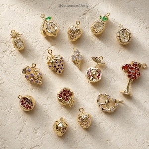 May include: An assortment of gold-coloured fruit and food charms, such as a pineapple, lemon, strawberry, grapes, apple, and pretzel. Each charm is adorned with sparkling gemstones, giving a luxurious and detailed look. The charms are displayed on a textured, off-white surface.