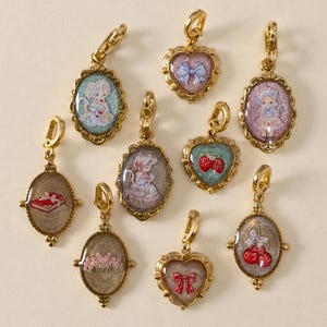 May include: A collection of nine gold-toned metal charms featuring oval and heart-shaped frames with glittery, colorful inserts. The designs include illustrations of figures, strawberries, cherries, a book, flowers, and bows. Each charm has a lobster clasp.